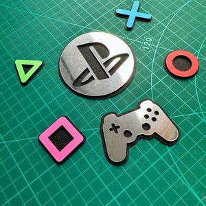 May include: A selection of PlayStation-themed cutouts, featuring a silver PlayStation logo, a controller, and coloured shapes: blue cross, red circle, green triangle, and pink square. The items are on a green grid surface.