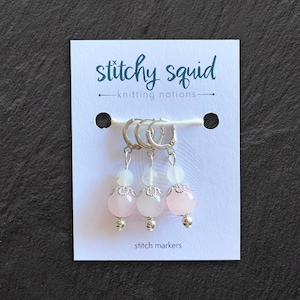 Op de afbeelding: Drie roze en witte steekmarkeerders met zilveren ringen. De markeerders bevinden zich op een witte kaart met de tekst "stitch markers" en "stitchysquid - knitting notions -" erop gedrukt.