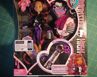 Op de afbeelding: Een Monster High Clawdeen Wolf pop in een doos. De pop heeft bruin haar en draagt een zwart jasje, een paarse en zwarte broek en een zwart topje. De doos heeft roze en zwarte accenten en toont het Monster High logo en de tekst "Clawdeen Wolf Daughter of The Werewolf".