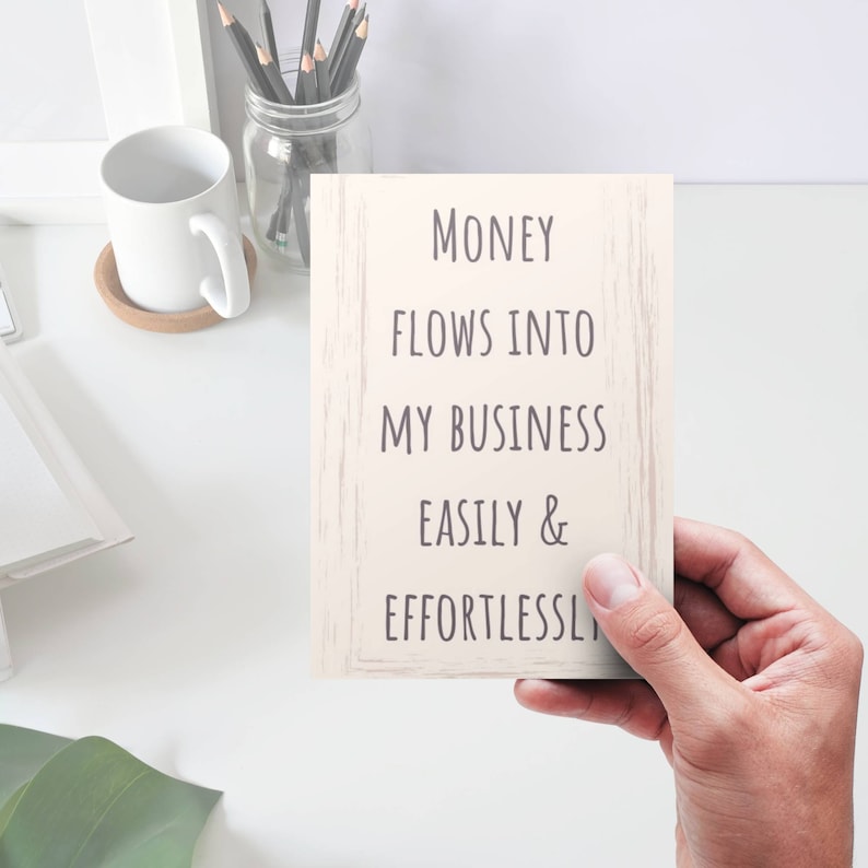 Puede incluir: Una tarjeta blanca con el texto "MONEY FLOWS INTO MY BUSINESS EASILY & EFFORTLESSLY" escrito en negro sobre un fondo de madera.