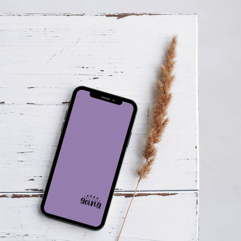 May include: A black smartphone with a lavender screen displaying the words "Keep Going" in a handwritten font. The phone rests on a distressed white wooden surface, accompanied by a dried, tan-colored plant stem.