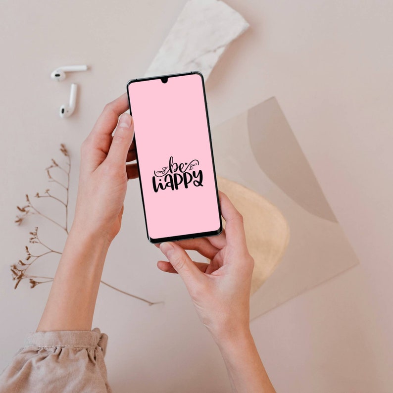 May include: A smartphone with a pink screen displaying the words "be happy" in black script is held by two hands. White wireless earbuds and a sprig of dried flowers are visible in the background. The phone has a black border.