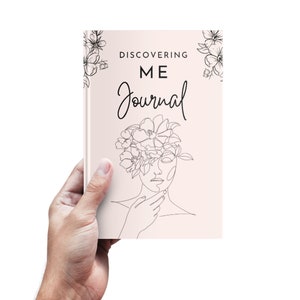 K&ouml;nnte beinhalten: Ein rosa Notizbuch mit dem Text "DISCOVERING ME Journal" und einer Strichzeichnung einer Frau mit Blumen auf dem Kopf.