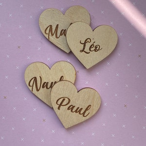 Peut inclure: Quatre ornements en bois en forme de cœur avec les noms "Ma", "Léo", "Nav" et "Paul" gravés dessus. Les ornements sont disposés sur un fond rose clair avec un motif blanc.