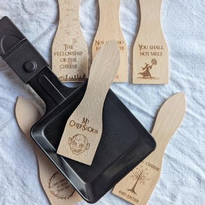 Peut inclure: Un appareil à raclette noir avec plusieurs spatules en bois. Les spatules portent des textes et des images gravés, dont "THE FELLOWSHIP OF THE CHEESE", "YOU SHALL NOT MELT" et "MY CHEESIOUS". Les spatules sont brun clair et le grill est noir.