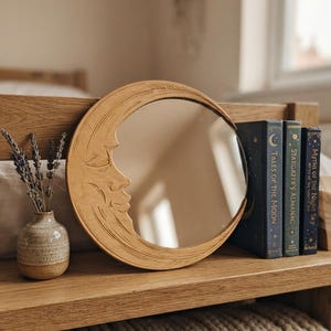 Peut inclure: Un miroir circulaire en bois avec un visage de lune croissante sculpté sur le côté gauche. À côté du miroir se trouvent trois livres intitulés Tales of the Moon, Stargazer's Almanac et Myths of the Night Sky, ainsi qu'un petit vase en céramique.
