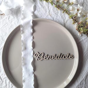 Peut inclure: Une assiette en céramique gris clair avec un marque-place en bois avec le nom "Bénédicte" en écriture cursive. Un ruban blanc à bord ondulé est posé sur l'assiette. Une branche de fleurs blanches et de feuilles vertes est en arrière-plan.