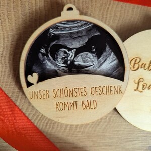 May include: A wooden Christmas ornament with a sonogram image of a baby. The ornament has a heart cutout and the German text "UNSER SCH&Ouml;NSTES GESCHENK KOMMT BALD" which translates to "Our most beautiful gift is coming soon."
