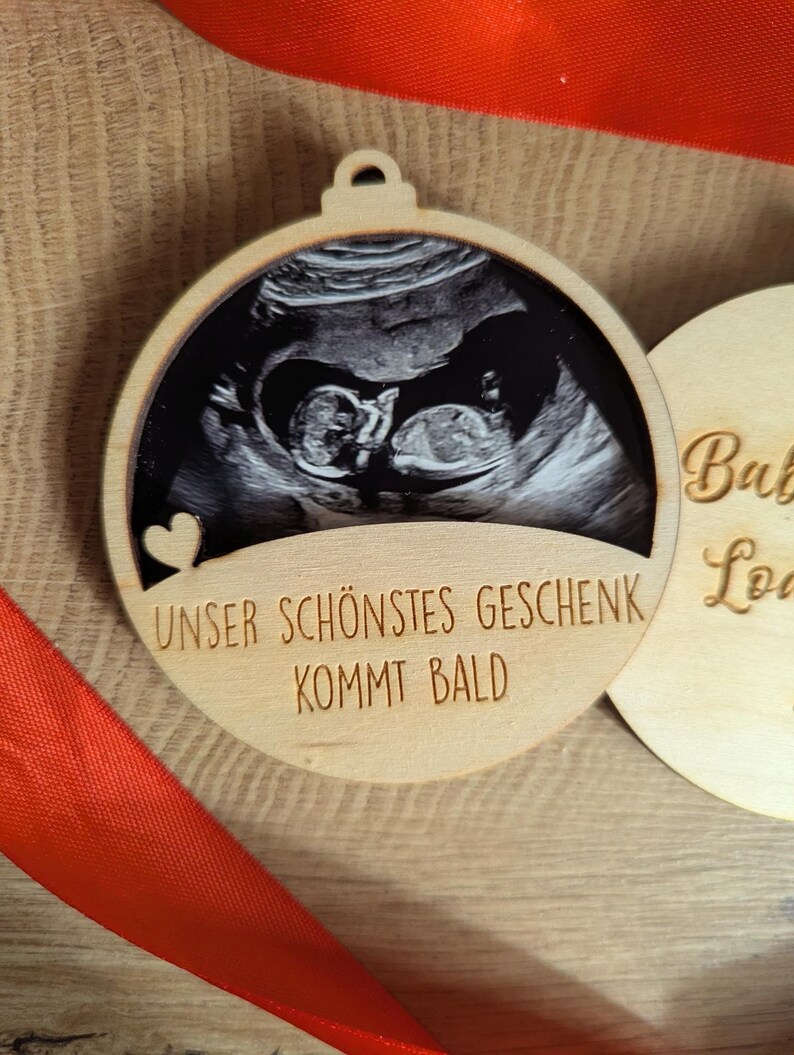 May include: A wooden Christmas ornament with a sonogram image of a baby. The ornament has a heart cutout and the German text "UNSER SCH&Ouml;NSTES GESCHENK KOMMT BALD" which translates to "Our most beautiful gift is coming soon."