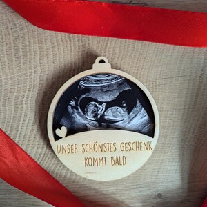 May include: A wooden ornament with a sonogram image of a baby. The ornament has a circular shape with a small loop at the top for hanging. The text "UNSER SCH&Ouml;NSTES GESCHENK KOMMT BALD" is engraved on the wood. A red ribbon is draped around the ornament.
