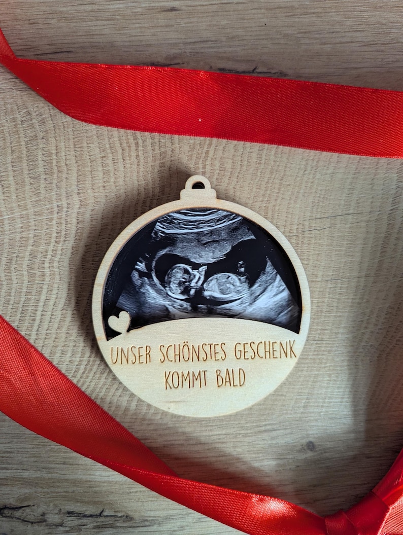 May include: A wooden ornament with a sonogram image of a baby. The ornament has a circular shape with a small loop at the top for hanging. The text "UNSER SCH&Ouml;NSTES GESCHENK KOMMT BALD" is engraved on the wood. A red ribbon is draped around the ornament.
