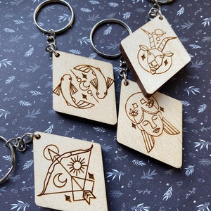 May include: Four wooden diamond-shaped keychains with zodiac signs engraved on them. The signs are Pisces, Capricorn, Gemini, and Sagittarius.