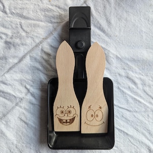 Könnte beinhalten: Zwei Holzspatel mit Cartoon-Gesichtern, eines mit einem zahnigen Grinsen und das andere mit einem fröhlichen Ausdruck, liegen in einem schwarzen Halter. Die Spatel sind hellbraun und der Halter ist schwarz. Der Hintergrund ist ein weißes, strukturiertes Gewebe.