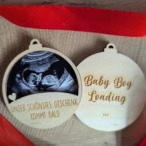 May include: Two wooden Christmas ornaments. One features a sonogram image and the text "Unser Sch&ouml;nstes Geschenk Kommt Bald". The other says "Baby Boy Loading" with ellipses. A red ribbon is draped across the ornaments.