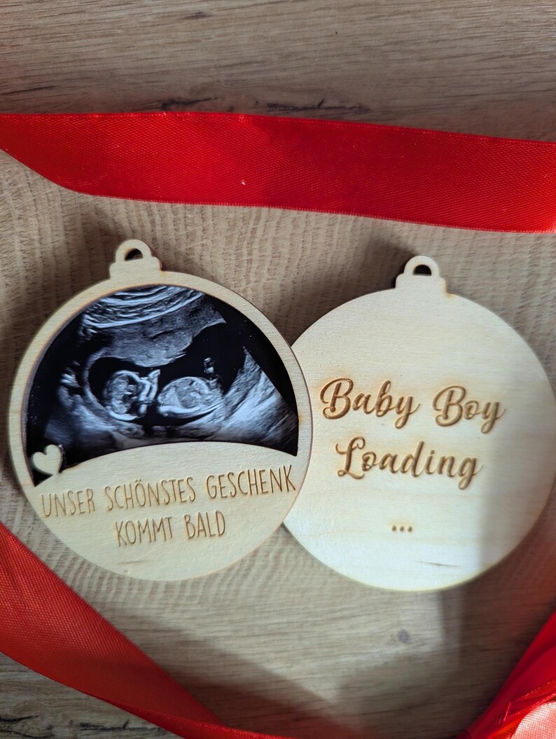 May include: Two wooden Christmas ornaments. One features a sonogram image and the text "Unser Sch&ouml;nstes Geschenk Kommt Bald". The other says "Baby Boy Loading" with ellipses. A red ribbon is draped across the ornaments.