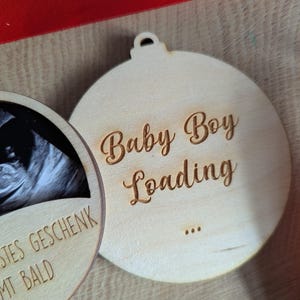 May include: Two wooden ornaments with a light wood finish. One features a baby ultrasound image, the other says "Baby Boy Loading" in script. A red ribbon is visible in the background. The ornaments are round with a small hole at the top for hanging.