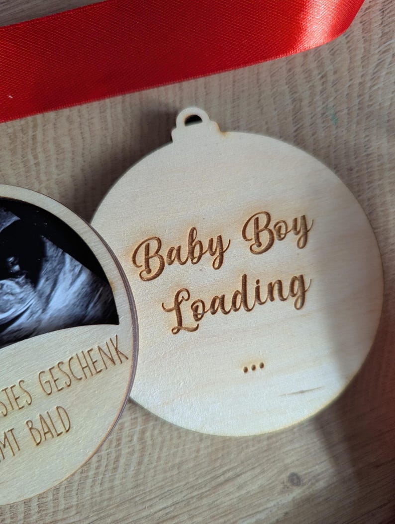 May include: Two wooden ornaments with a light wood finish. One features a baby ultrasound image, the other says "Baby Boy Loading" in script. A red ribbon is visible in the background. The ornaments are round with a small hole at the top for hanging.