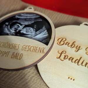 May include: Two wooden ornaments with a light brown finish. One features a black and white ultrasound image and the German text "SCH&Ouml;NSTES GESCHENK KOMMT BALD". The other ornament has the words "Baby is Loading" in a script font.