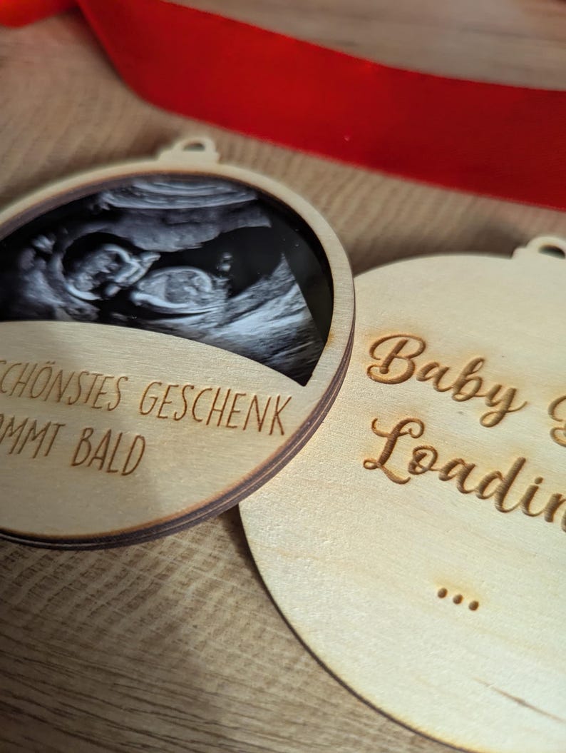 May include: Two wooden ornaments with a light brown finish. One features a black and white ultrasound image and the German text "SCH&Ouml;NSTES GESCHENK KOMMT BALD". The other ornament has the words "Baby is Loading" in a script font.