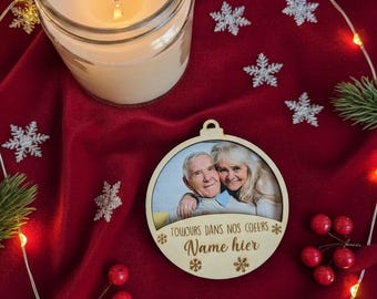 May include: A lit candle in a glass jar sits on a red velvet surface, surrounded by snowflake ornaments, red berries, and a string of lights. A wooden ornament with a photo and text is also present.