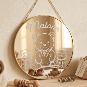 Peut inclure: Un miroir rond à cadre doré suspendu par une chaîne dorée. Le miroir présente un motif blanc gravé représentant un ours en peluche, deux piles de cubes et le prénom Malonn. Les cubes affichent le texte 08, Juillet, 2024, 3.140 kg et 49.5cm.