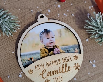 May include: Wooden Christmas ornament with a photo of a baby. The ornament has the text "MON PREMIER NOËL Camille 2025" engraved on it. The ornament is round with a hole at the top for hanging. Decorative elements include snowflakes and a sprig of evergreen.