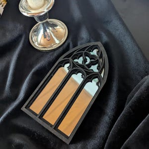 Può includere: Uno specchio nero in stile gotico con tre sezioni rettangolari a specchio. Lo specchio ha una parte superiore ad arco con intagli decorativi. Lo specchio è esposto su una superficie di velluto nero, con un candelabro d'argento e una candela sullo sfondo.