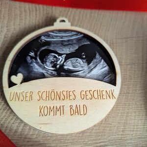 May include: Wooden Christmas ornament with a sonogram image of a baby. The ornament has the text "UNSER SCH&Ouml;NSTES GESCHENK KOMMT BALD" and a small heart. A red ribbon is visible in the background.