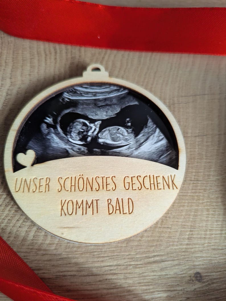 May include: Wooden Christmas ornament with a sonogram image of a baby. The ornament has the text "UNSER SCH&Ouml;NSTES GESCHENK KOMMT BALD" and a small heart. A red ribbon is visible in the background.