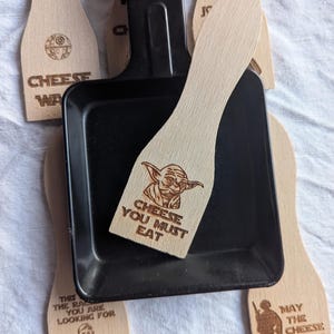 Könnte beinhalten: Ein schwarzes Raclette-Grillpfännchen mit mehreren Holz-Käsespateln. Die Spatel haben eingravierte Star Wars-Motive und Sätze wie "Cheese You Must Eat" und "May the Cheese Be With You."