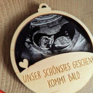May include: A wooden ornament with a sonogram image of a baby. The ornament has a heart cutout and the text "UNSER SCH&Ouml;NSTES GESCHENK KOMMT BALD" engraved on it. The ornament is round with a small loop at the top.