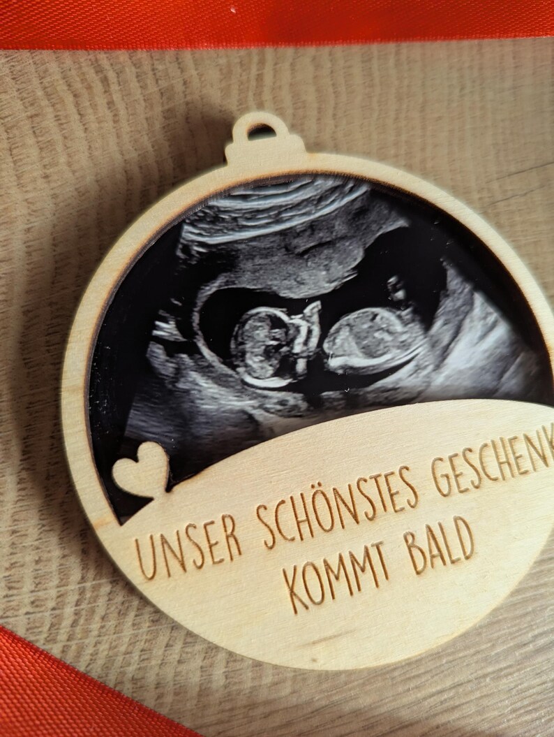 May include: A wooden ornament with a sonogram image of a baby. The ornament has a heart cutout and the text "UNSER SCH&Ouml;NSTES GESCHENK KOMMT BALD" engraved on it. The ornament is round with a small loop at the top.