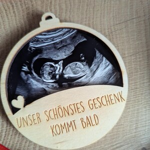 May include: A wooden ornament with a sonogram image of a baby. The ornament has a heart cutout and the German text "UNSER SCH&Ouml;NSTES GESCHENK KOMMT BALD" which translates to "Our most beautiful gift is coming soon."