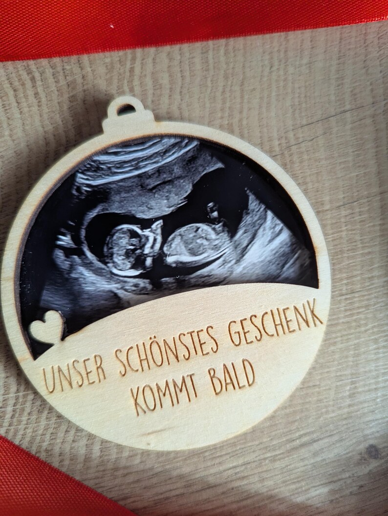 May include: A wooden ornament with a sonogram image of a baby. The ornament has a heart cutout and the German text "UNSER SCH&Ouml;NSTES GESCHENK KOMMT BALD" which translates to "Our most beautiful gift is coming soon."