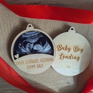 May include: Two wooden ornaments with a red ribbon. One ornament features a sonogram image and the text "Unser Sch&ouml;nstes Geschenk Kommt Bald". The other ornament says "Baby Boy Loading".