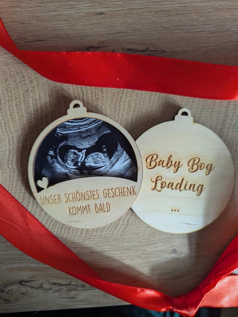 May include: Two wooden ornaments with a red ribbon. One ornament features a sonogram image and the text "Unser Sch&ouml;nstes Geschenk Kommt Bald". The other ornament says "Baby Boy Loading".