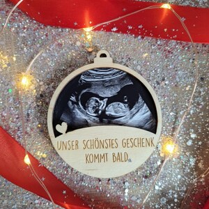 May include: Wooden ornament with a sonogram image, a heart, and the text "UNSER SCH&Ouml;NSTES GESCHENK KOMMT BALD." The ornament is surrounded by a red ribbon and fairy lights, set against a glittery background.