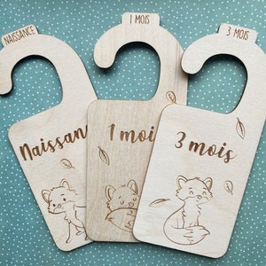 Peut inclure: Trois cintres en bois avec des illustrations de renards et du texte en français. Les cintres sont étiquetés "Naissance", "1 mois" et "3 mois".