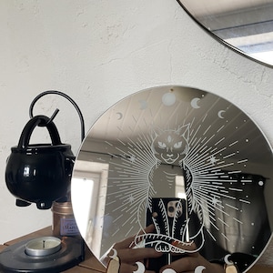 Peut inclure: Un miroir rond argenté avec un chat noir et des phases de lune gravées sur la surface. Le chat a trois yeux et est entouré d'un motif de soleil.