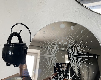 May include: A round, silver-toned mirror with a black cat and moon phases etched into the surface. The cat has three eyes and is surrounded by a sunburst design.