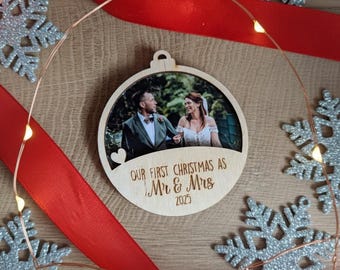 May include: Circular wooden Christmas ornament featuring a couple's photo, the text "OUR FIRST CHRISTMAS AS Mr & Mrs 2025", and a heart. Decorated with silver glitter snowflakes, a red ribbon, and fairy lights.