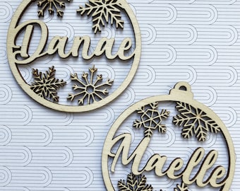 May include: Two wooden Christmas ornaments with the names "Danae" and "Maelle" cut out in cursive script. Each ornament is a circle with snowflake cutouts and a loop for hanging. The ornaments are light brown.