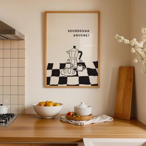 Op de afbeelding: Een ingelijste kunstprint met de tekst "SOURDOUGH ANYONE?" toont een zwart-wit illustratie van een koffiezetapparaat, kopjes en brood op een dambordpatroon. De print zit in een licht houten frame, weergegeven in een keuken.