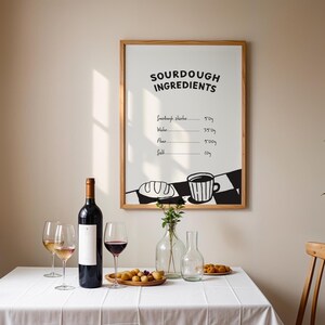 Op de afbeelding: Een eettafel met een wit tafelkleed, wijnglazen en een fles rode wijn. Een ingelijste print aan de muur geeft de ingrediënten voor zuurdesem weer. Een houten bord bevat kleine, ronde etenswaren. Een vaas bevat een paar groene stelen.