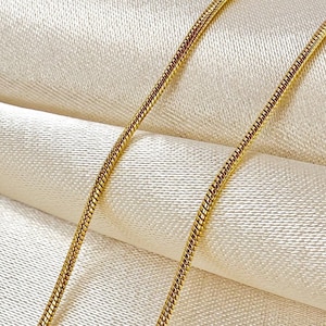 Puede incluir: Una cadena de cuello flexible y delgada de tono dorado, tipo serpiente, que presenta un perfil redondeado y liso con un patrón continuo de eslabones entrelazados. La superficie metálica refleja la luz, resaltando la estructura cilíndrica uniforme de la pieza.
