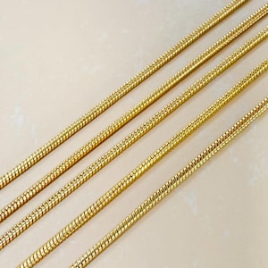 May include: Four parallel gold-toned snake chains arranged diagonally across the frame. Each chain features a smooth, flexible, and tightly linked cylindrical design with a reflective metallic finish that catches the light along its length.