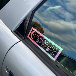 May include: A holographic sticker with the text "LOST NIGHTS" and the Japanese text "失われた夜" in pink and blue lettering on a silver car window.