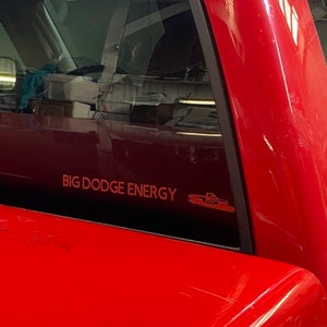 Può includere: Un pick-up rosso con un adesivo sul lunotto posteriore che recita "BIG DODGE ENERGY" con una piccola icona di camion accanto.