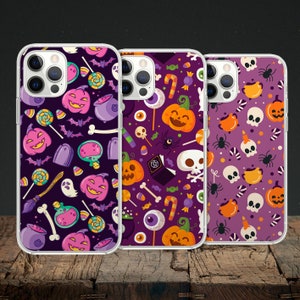 Op de afbeelding: Drie transparante telefoonhoesjes met kleurrijke Halloween-ontwerpen. Elk hoesje heeft een ander patroon van pompoenen, snoep, schedels en vleermuizen op een paarse achtergrond.