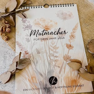 Könnte beinhalten: Ein Spiral-Kalender für 2026 mit einem Aquarell-Blumenmuster in sanften Pfirsich-, Lavendel- und Grüntönen. Der Kalender trägt die Aufschrift "Mutmacher" und "FÜR DEIN JAHR 2026". Unten steht "EIN KALENDER DER MUT & HOFFNUNG SCHENKT".